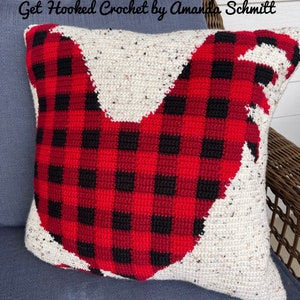 May include: A red and black plaid crocheted pillow with a white crocheted rooster design. The pillow is on a blue couch with a wicker chair in the background. The text "Get Hooked Crochet by Amanda Schmitt" is visible at the top of the image.