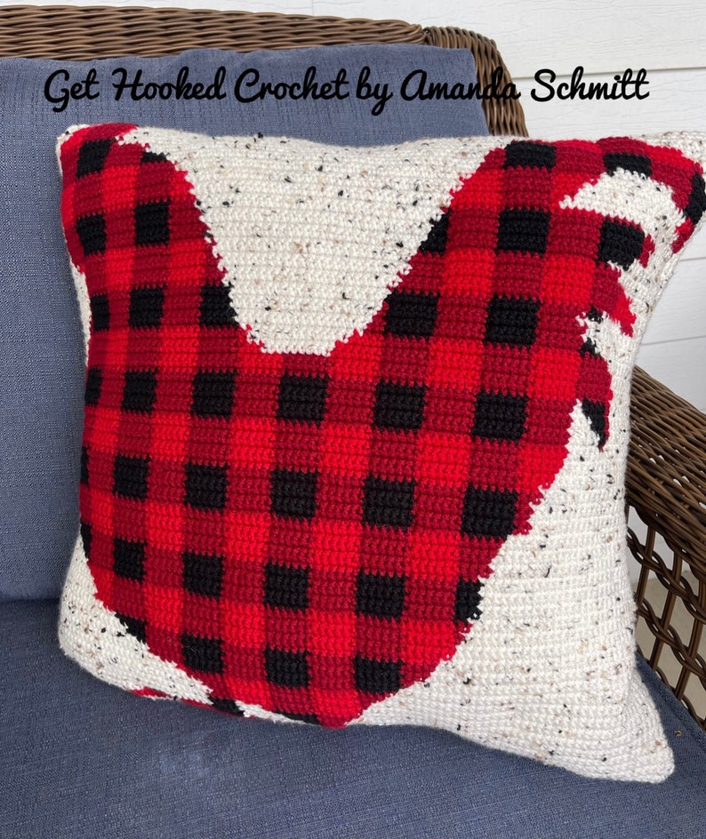 May include: A red and black plaid crocheted pillow with a white crocheted rooster design. The pillow is on a blue couch with a wicker chair in the background. The text "Get Hooked Crochet by Amanda Schmitt" is visible at the top of the image.