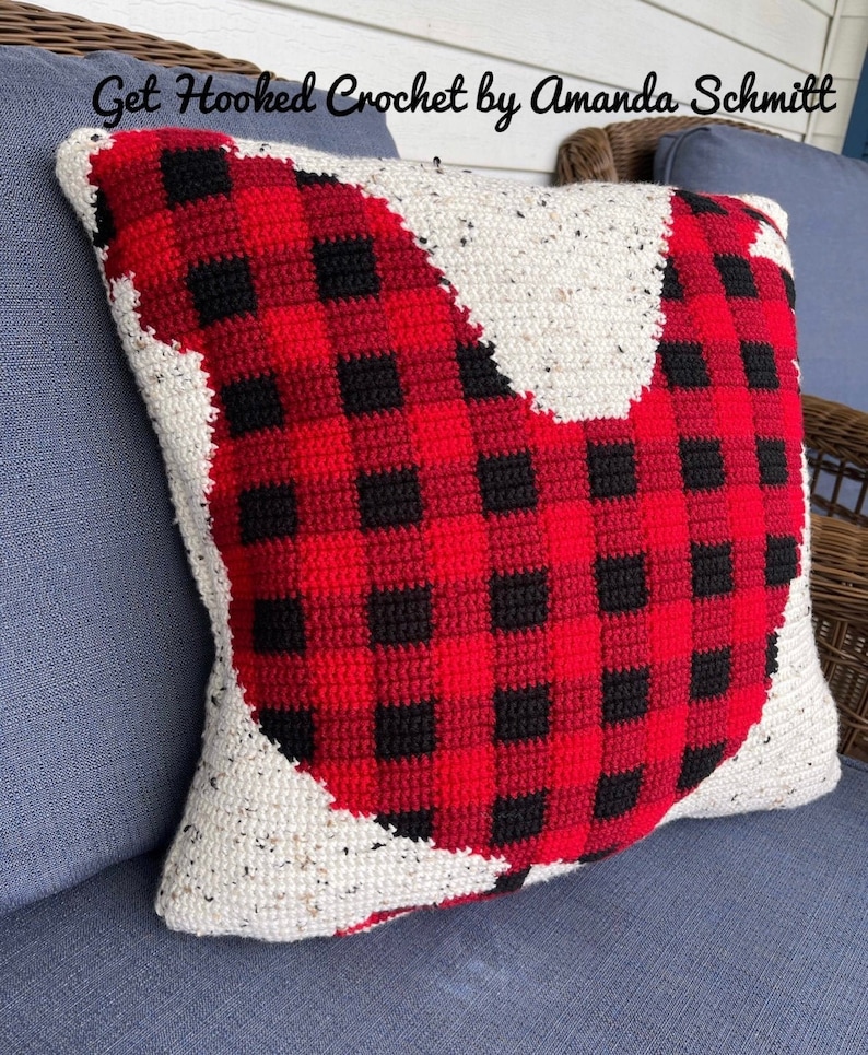 May include: A red and black plaid crocheted pillow in the shape of a chicken. The pillow is sitting on a blue couch.