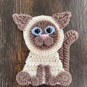 May include: A crocheted Siamese cat with brown and white fur, blue eyes, and a pink nose. The cat is sitting on a wooden surface.