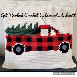 May include: Crocheted pillow featuring a red truck with a green Christmas tree in the bed. The truck is decorated with a black and red buffalo plaid pattern. The pillow is white with a crocheted texture. The text "Get Hooked Crochet by Amanda Schmitt" is at the top of the pillow.