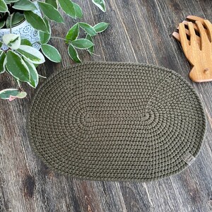 May include: An oval-shaped, olive green crocheted placemat featuring a dense, uniform stitch pattern. A small, rectangular fabric tag is attached to the lower right edge of the placemat, which is positioned on a dark, wood-grain surface.