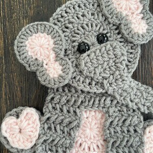 May include: A gray crocheted elephant with pink ears and feet. The elephant is sitting on a wooden surface. The text "Get Hooked Crochet by Amanda Schmitt" is at the top of the image. The text "soft Pink" is at the bottom of the image.