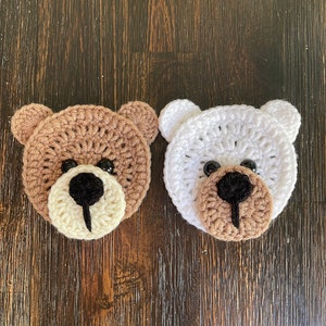 May include: Two crocheted bear faces, one brown and one white, with black eyes and noses. The bears are on a dark brown wooden surface.