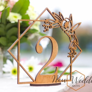 May include: Wooden table number with a geometric design and floral accents. The number two is displayed in the centre of the design. The table number is made of natural wood and has a rustic look. The text "New Wedding Signs" is visible at the bottom of the image.