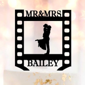 May include: A wedding cake with white frosting and gold leaf accents, topped with a black film strip-shaped cake topper. The topper reads "MR & MRS" above a silhouette of a couple and "BAILEY" below.