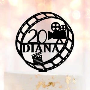 May include: A black cake topper with the text "20 DIANA" and film reel and camera graphics. The topper sits atop a white cake decorated with gold leaf and pink roses.