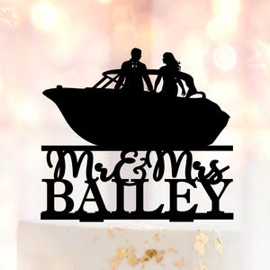 Puede incluir: Una tarta de boda blanca con decoración de pan de oro, coronada con una silueta negra de una pareja en un barco. La tarta está decorada con rosas rosas y una decoración para tarta que dice "Mr. & Mrs. Bailey."