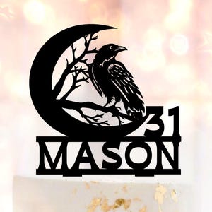 May include: A white cake with gold leaf accents and pink roses. A black cake topper features a raven perched on a branch within a crescent moon, with the name "MASON" and the number "31".
