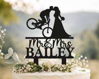 May include: A black cake topper featuring a silhouette of a person on a mountain bike kissing a person in a wedding dress. Below the figures, the text reads Mr & Mrs BAILEY in a combination of script and block lettering with two support stakes.