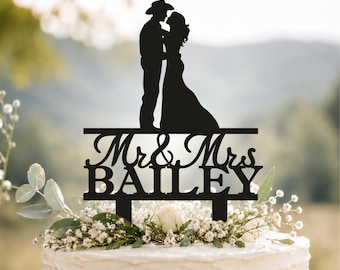 May include: A black cake topper featuring a silhouette of a couple, with the person on the left wearing a cowboy hat. Below the couple, the text reads Mr & Mrs BAILEY in a combination of cursive and block lettering, supported by two stakes.