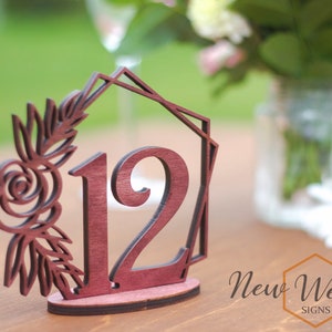 Op de afbeelding: Houten tafelnummer met een bloemmotief en het nummer 12. De tafelnummer is donker roodbruin van kleur. Het bloemmotief is uit het hout gesneden en is lichtbruin van kleur. Het nummer 12 is ook uit het hout gesneden en is lichtbruin van kleur. De tafelnummer staat op een houten tafel. De tekst "New Wedding Signs" is zichtbaar in de rechterbenedenhoek van de afbeelding.