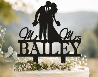 May include: A black cake topper featuring a silhouette of a couple kissing, with the text "Mr & Mrs" in cursive script above the block-lettered surname "BAILEY". The topper has two prongs at the base for insertion into a cake surface.