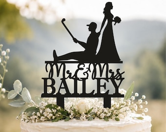 Op de afbeelding: Een zwarte taarttopper met een silhouet van een persoon met een pet die een golfclub vasthoudt en een persoon in een lange jurk met een boeket. De tekst Mr & Mrs BAILEY staat in een combinatie van script en blokletters onder de figuren.