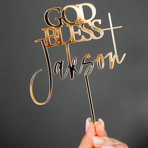 May include: Gold-colored cake topper with the words "God Bless Jackson" in a cursive font.