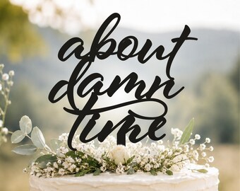 May include: A black cake topper featuring the phrase about damn time in a cursive script font. The topper is positioned on top of a white frosted cake decorated with a cluster of small white flowers and green leaves, set against a blurred background.