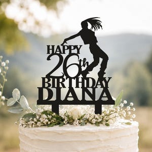 May include: A black cake topper featuring a silhouette of a person dancing above the text HAPPY 26th BIRTHDAY DIANA. The topper has two prongs at the base and is positioned on top of a white frosted cake decorated with small white flowers.