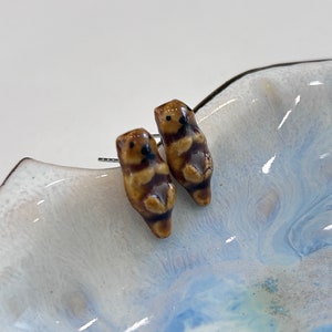 Könnte beinhalten: Ein Paar braune Keramik-Otter-Ohrringe. Die Otter schauen sich an und haben schwarze Augen. Die Ohrringe befinden sich auf einem weißen und blauen Hintergrund.