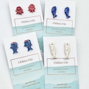 May include: Four pairs of ceramic stud earrings on display. The earrings feature sea creatures: red octopuses, blue dolphins, blue fish, and white whales. Each pair is mounted on a card with the text "Chihiro Uki porcelain jewelry / ceramics".