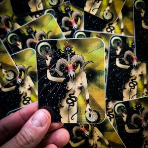 May include: A sticker featuring a colorful illustration of a woman with a ram skull head, holding a crescent moon in one hand and a dagger in the other. The background is a yellow and black gradient with stars.