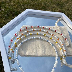 May include: Four white beaded necklaces with colorful accents. The necklaces are arranged in a rainbow pattern, with red, orange, yellow, and blue beads. The necklaces are displayed on a white surface with a white cloth bag.