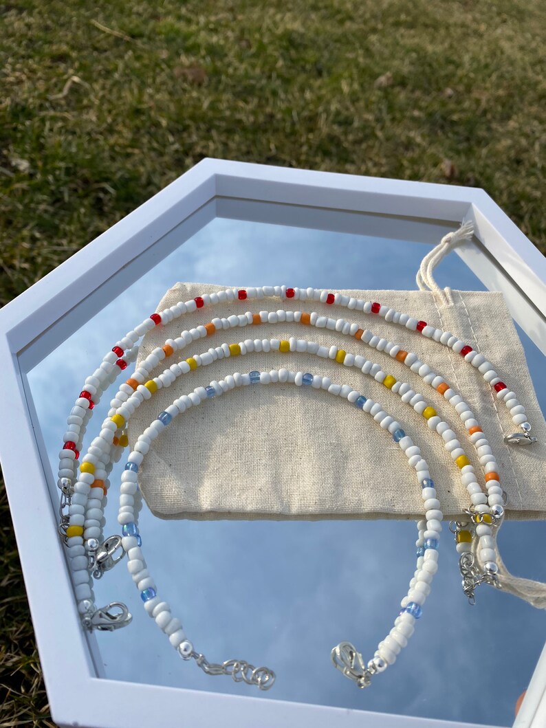 May include: Four white beaded necklaces with colorful accents. The necklaces are arranged in a rainbow pattern, with red, orange, yellow, and blue beads. The necklaces are displayed on a white surface with a white cloth bag.