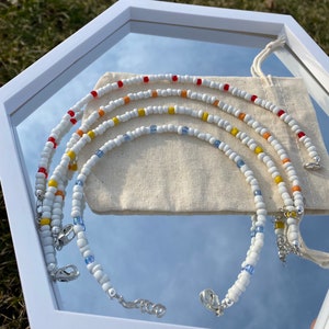 May include: Four white beaded necklaces with colorful accents. The necklaces are layered and feature red, orange, yellow, and blue beads. The necklaces are displayed on a white background.