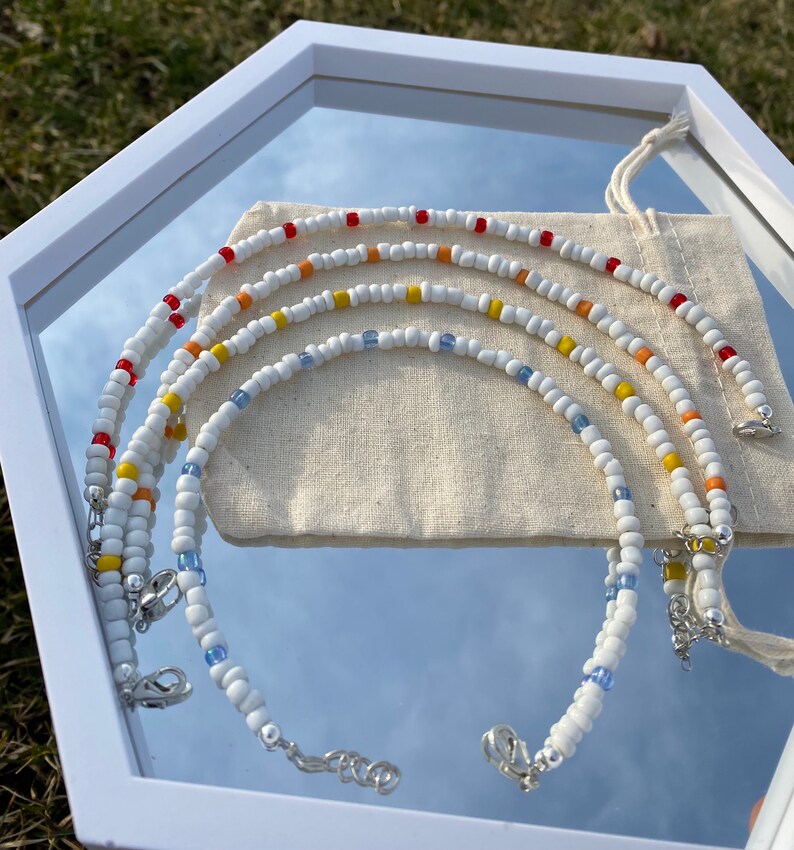 May include: Four white beaded necklaces with colorful accents. The necklaces are layered and feature red, orange, yellow, and blue beads. The necklaces are displayed on a white background.