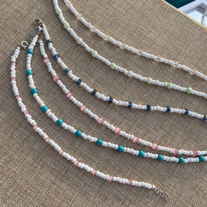 May include: Five beaded necklaces with white beads and different colored accent beads. The necklaces are arranged in a fan shape on a brown textured surface.
