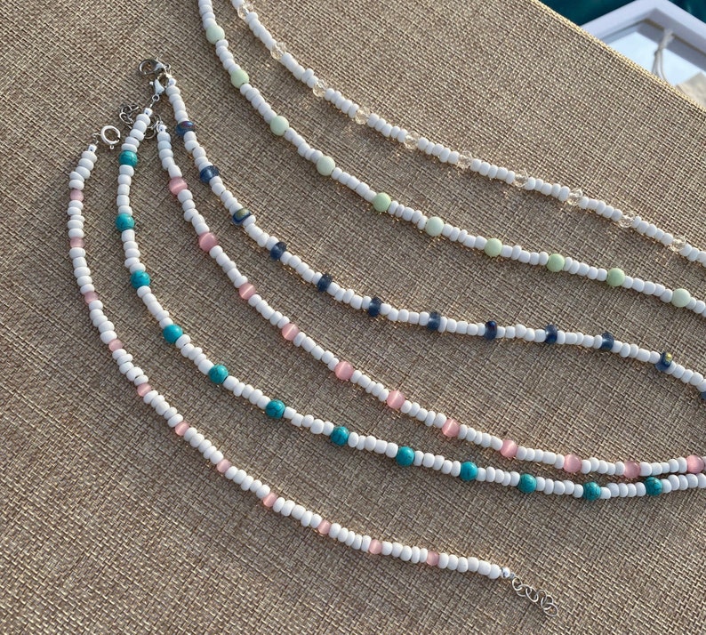 May include: Five beaded necklaces with white beads and different colored accent beads. The necklaces are arranged in a fan shape on a brown textured surface.