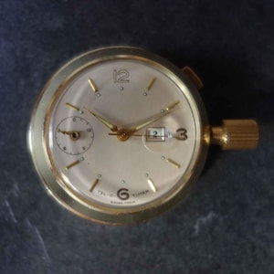 Könnte beinhalten: Eine goldfarbene Taschenuhr mit weißem Zifferblatt und schwarzen Zeigern. Die Uhr hat ein kleines Hilfszifferblatt auf 3 Uhr und ein Datumsfenster auf 4 Uhr. Das Zifferblatt der Uhr trägt außerdem die Aufschrift "Tel-O-Timer" und "Swiss Made".