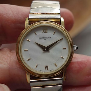 May include: A gold-tone Wittnauer wristwatch with a white face and black hands. The watch has a two-tone band with a gold-tone top and a silver-tone bottom.