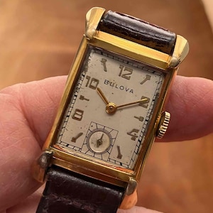 May include: Vintage Bulova watch with a rectangular gold-tone case and a cream-coloured face. The watch features black numerals, gold hands, and a subdial. The watch has a dark brown leather strap.