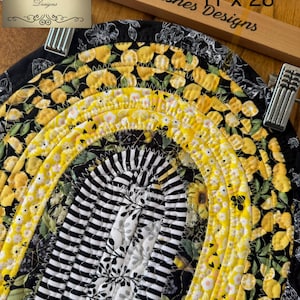 May include: Oval-shaped quilted placemat with a vibrant design. The placemat features a combination of black and white striped fabric, yellow floral patterns, and a black floral border. The dimensions are 11 x 28 inches.