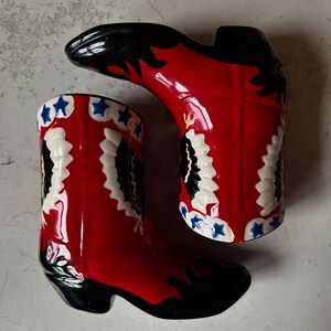 May include: A pair of ceramic cowboy boots. The boots are primarily red with black accents on the toe and top. White and black decorative patterns adorn the sides, with blue star details. The boots are designed to stand upright.