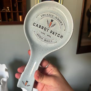 May include: A light blue ceramic spoon rest with dark speckles. The center features black text reading LOCALLY GROWN CARROT PATCH EST. 1976 OPEN DAILY, with a small orange carrot graphic. A smaller carrot graphic is located on the handle.