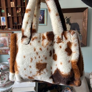 Puede incluir: Un bolso rectangular de piel sint&eacute;tica con estampado de vaca. El bolso tiene una base blanca con manchas marrones y ribetes y asas marrones. Las asas son gruesas y afelpadas, a juego con la textura del bolso. El bolso est&aacute; sostenido por una mano.