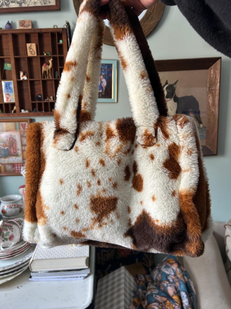Puede incluir: Un bolso rectangular de piel sint&eacute;tica con estampado de vaca. El bolso tiene una base blanca con manchas marrones y ribetes y asas marrones. Las asas son gruesas y afelpadas, a juego con la textura del bolso. El bolso est&aacute; sostenido por una mano.