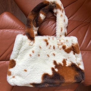 Puede incluir: Un bolso tote esponjoso con un estampado de piel de vaca. El bolso presenta una base blanca con manchas marrones y marr&oacute;n oscuro, y asas a juego. El bolso est&aacute; hecho de un material suave y afelpado.