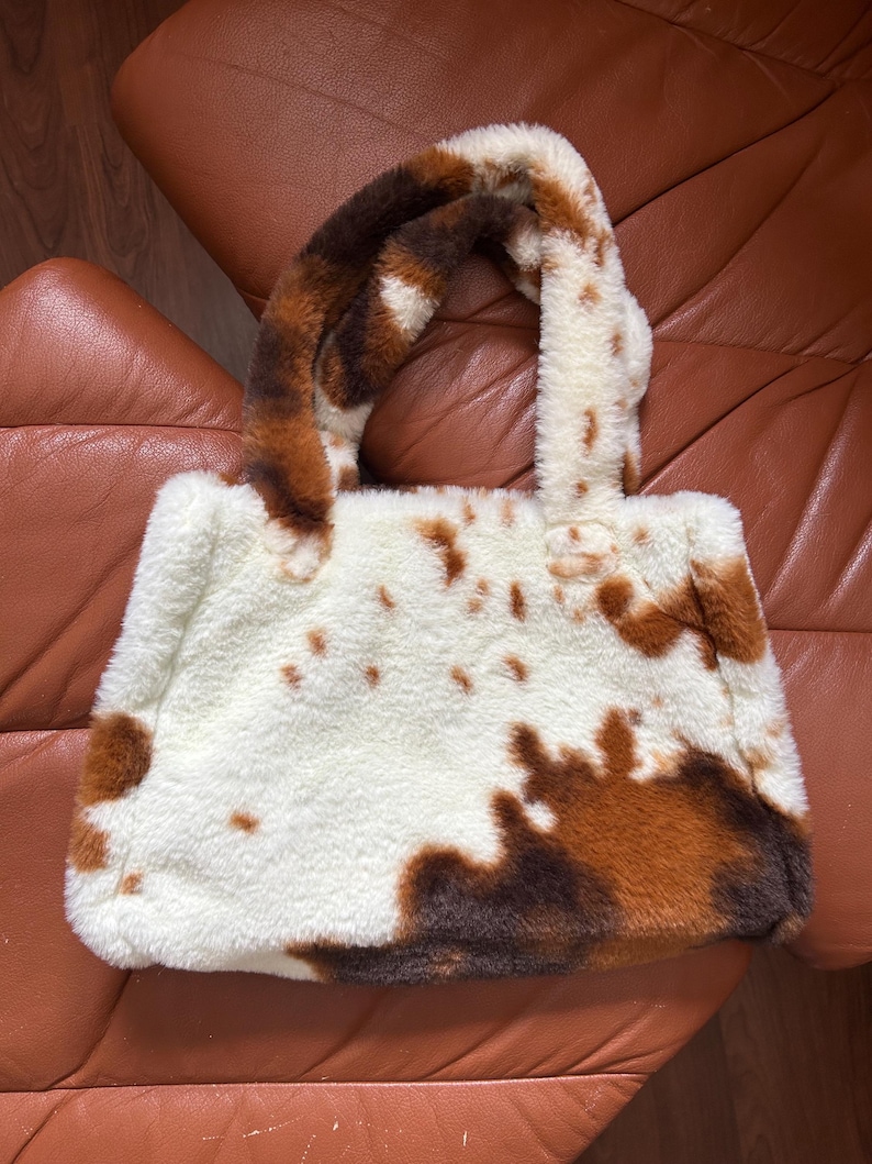 Puede incluir: Un bolso tote esponjoso con un estampado de piel de vaca. El bolso presenta una base blanca con manchas marrones y marr&oacute;n oscuro, y asas a juego. El bolso est&aacute; hecho de un material suave y afelpado.