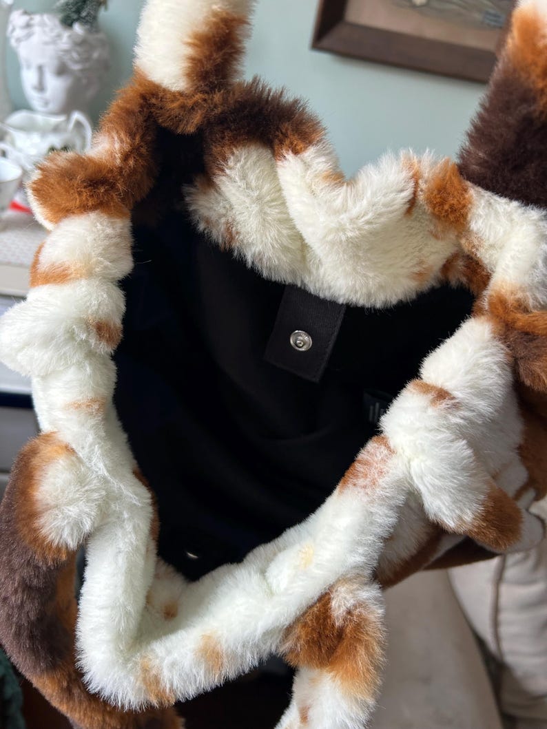 Puede incluir: Bolso tote de piel sint&eacute;tica con un patr&oacute;n marr&oacute;n, crema y blanco. El bolso tiene una textura suave y esponjosa y un interior negro. Una peque&ntilde;a etiqueta negra con un cierre a presi&oacute;n es visible.