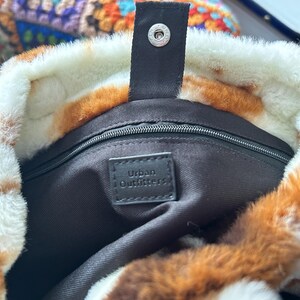 Puede incluir: Primer plano de un bolso tote de piel sint&eacute;tica con un estampado de manchas marrones y blancas. El bolso tiene una correa negra con un cierre de bot&oacute;n a presi&oacute;n plateado. El interior es negro con una etiqueta "Urban Outfitters."