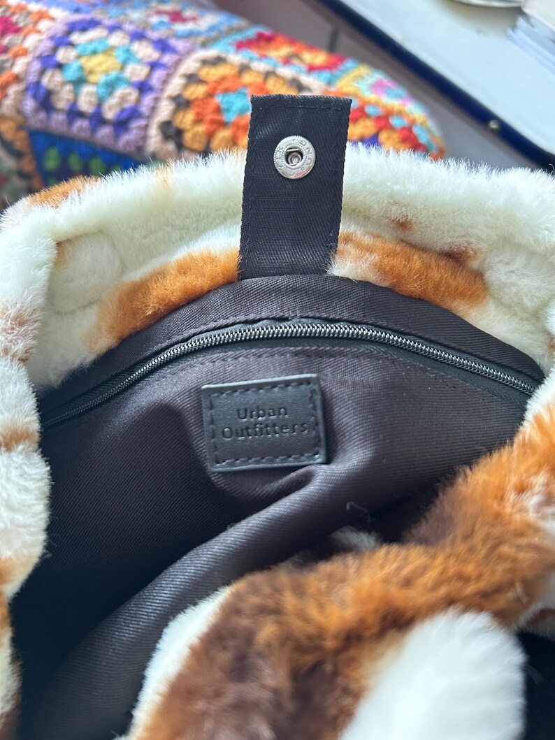 Puede incluir: Primer plano de un bolso tote de piel sint&eacute;tica con un estampado de manchas marrones y blancas. El bolso tiene una correa negra con un cierre de bot&oacute;n a presi&oacute;n plateado. El interior es negro con una etiqueta "Urban Outfitters."