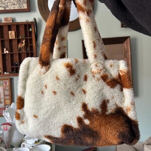 Puede incluir: Un bolso de mano de piel sint&eacute;tica con estampado de vaca. El bolso tiene una base blanca con manchas marrones y asas marrones y blancas a juego. El bolso es rectangular con una textura suave y afelpada.