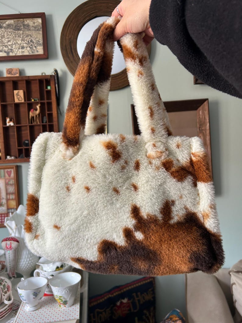 Puede incluir: Un bolso de mano de piel sint&eacute;tica con estampado de vaca. El bolso tiene una base blanca con manchas marrones y asas marrones y blancas a juego. El bolso es rectangular con una textura suave y afelpada.