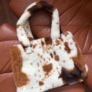 Puede incluir: Un bolso tote de piel sint&eacute;tica con dise&ntilde;o de estampado de vaca. El bolso presenta una base blanca con manchas marrones y un asa a juego. El bolso es rectangular y tiene una textura suave y afelpada.