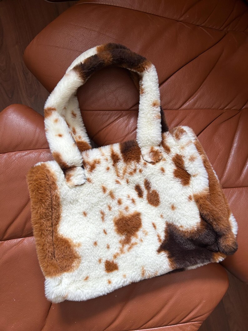 Puede incluir: Un bolso tote de piel sint&eacute;tica con dise&ntilde;o de estampado de vaca. El bolso presenta una base blanca con manchas marrones y un asa a juego. El bolso es rectangular y tiene una textura suave y afelpada.