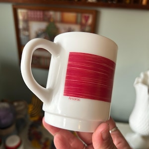 May include: A white mug with a smooth finish and a curved handle. The side of the mug features a rectangular graphic composed of horizontal red lines. The word Arcoroc is printed in small red letters at the bottom of the red graphic design.