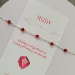Könnte beinhalten: Ein filigranes Armband aus Silberkette mit kleinen, leuchtend roten Rubinperlen. Das Armband wird auf einer weißen Karte mit dem Wort "RUBY" und den Sätzen "Vitalität-Energie-Leidenschaft" und "Bedingungslose Liebe-Reichtum" präsentiert.