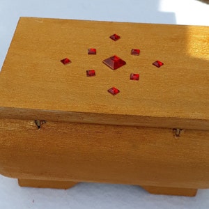 Könnte beinhalten: Eine Holzkiste mit goldener Oberfläche und einem dekorativen Design aus roten Strasssteinen. Die Kiste hat einen Deckel mit Scharnier und einen abgerundeten Boden.